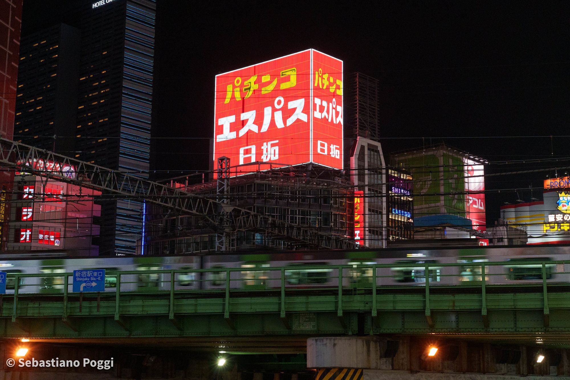 Picture of a Yamanote Line train zooming by on the Shinjuku rail bridge at night, with Kabukicho in the background